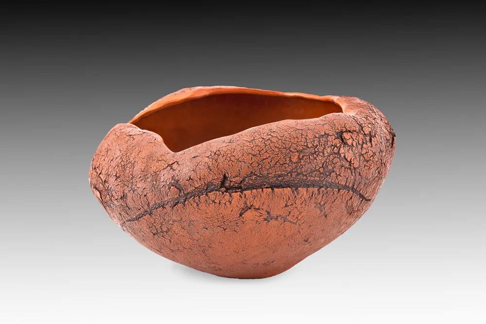 A rough-textured, hand-built terracotta bowl with a cracked, organic surface photographed against a plain gray gradient background